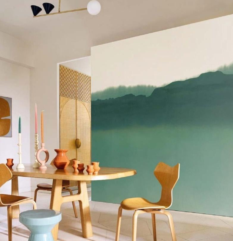 A wall mural featuring a minimalist landscape with green and teal mountains under a pale sky, shown installed in a dining room alongside a wooden table and chairs. The right side of the image is a dark panel with product details for the 'Pittore' mural, including dimensions and roll size.